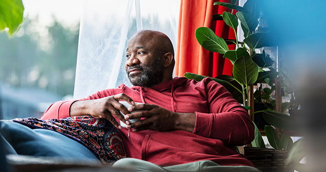 A middle aged man sits on his couch with coffee looking out the window